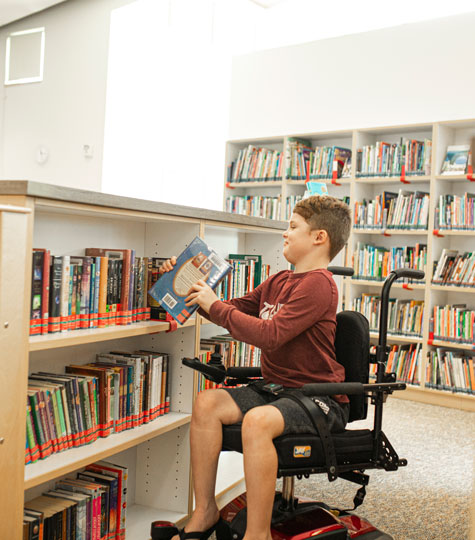 Boy with Duchenne reaching from wheelchair for book on low library shelf